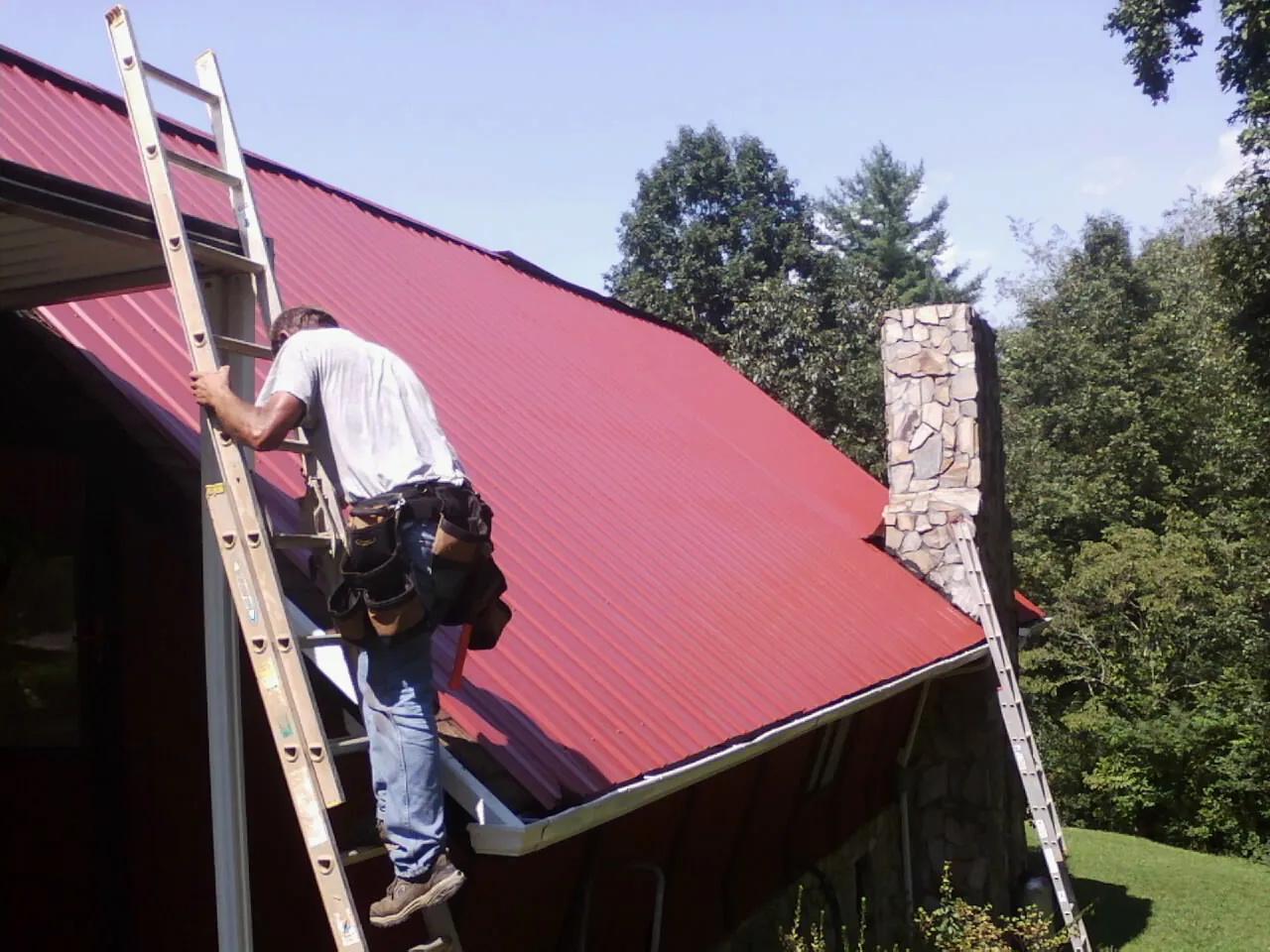 Roofer on a ladder beside a red metal roof and stone chimney — Soffit & Fascia Repair by Roofing Brothers in Baring